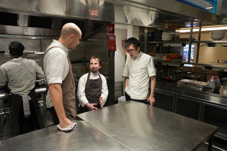 Robbie and Darren are seen wearing white chef's jackets, standing in the kitchen talking to a tall man in an apron.