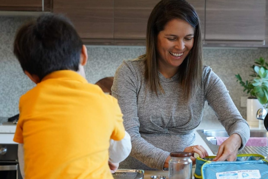 A pregnant woman in a grey dress in a kitchen making lunch for a boy in a yellow school uniform.