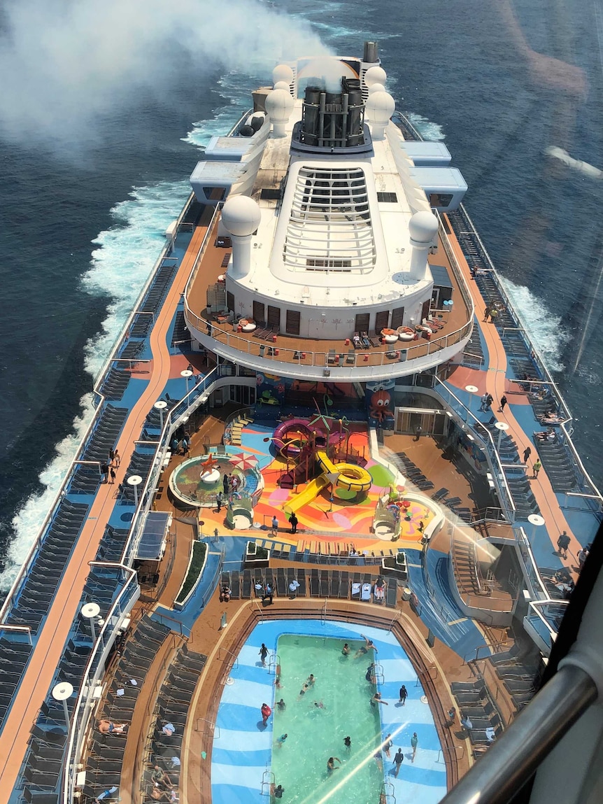 A cruise ship sails while people swim in pools or lay on the deck.