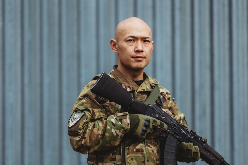 A bald man in military uniform poses with an assault rifle