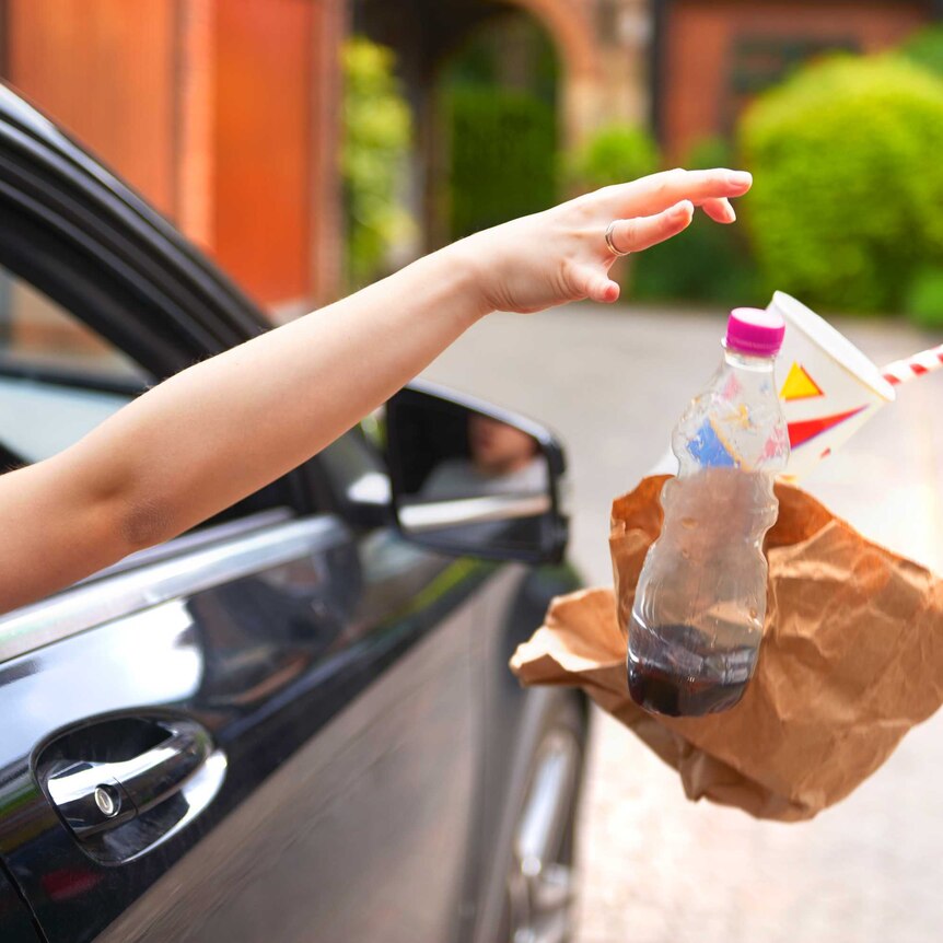 An arm and hand shown throwing take-away drink containers and rubbish out of a car window.