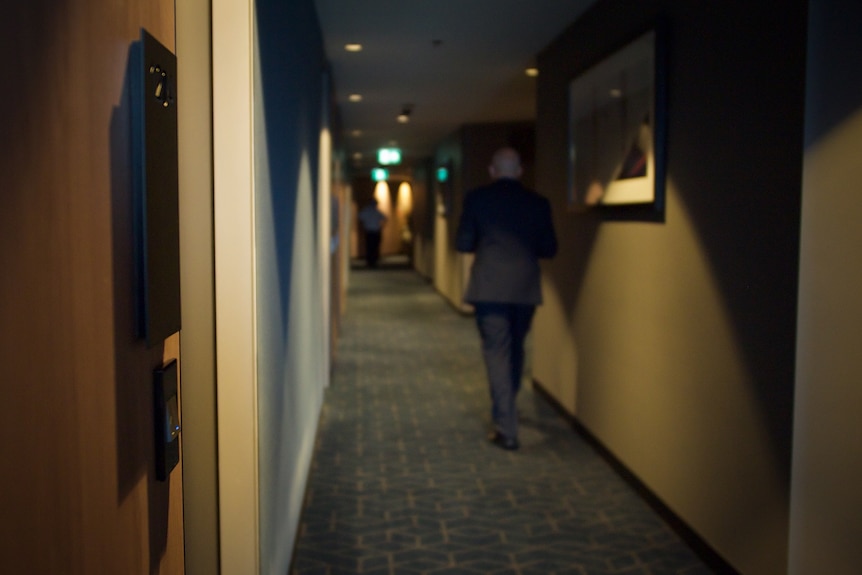 A photograph of a hotel corridor focuses on a room door as a bald man in a suit walks away. 