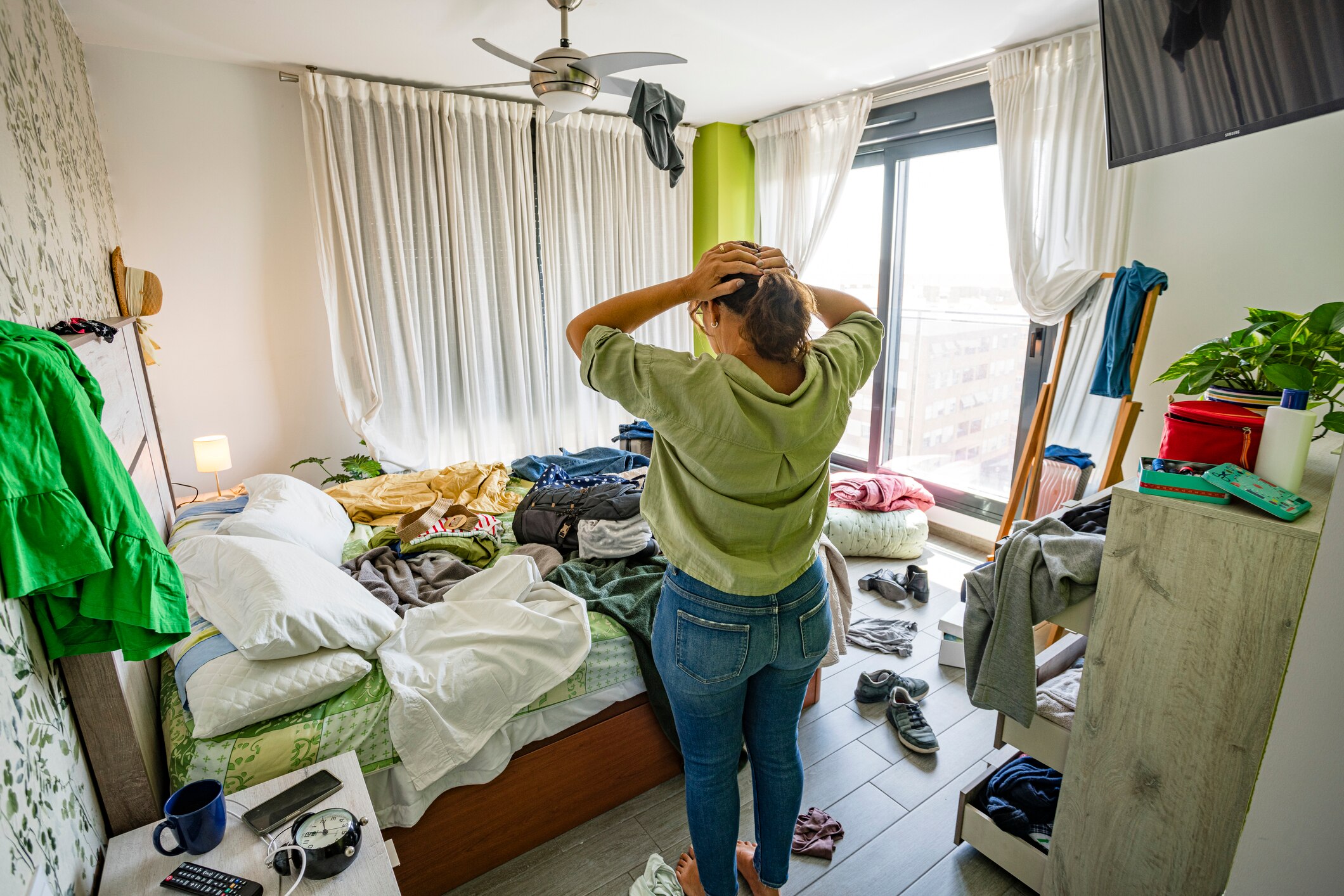 A woman holds her head as she looks at her messy bedroom