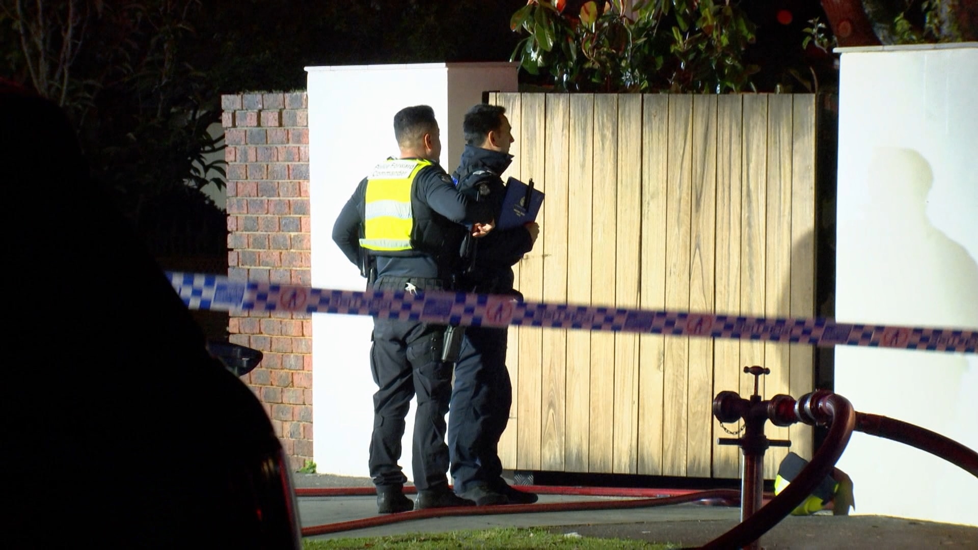 Two police officers stand in front of a wooden fence with police tape in the foreground.