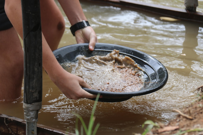 A person crouches with a gold pan in their hands above a muddy river.
