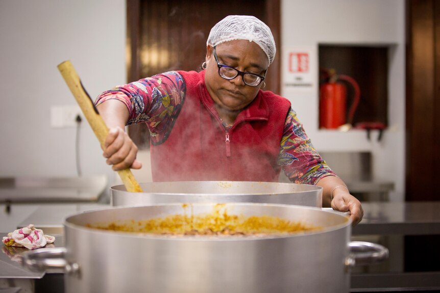 Woman wearing a hair net stirs a large pot of steaming food.
