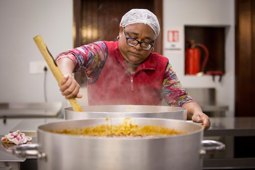 A woman wearing a hair net stirs a large amount of steamed food.