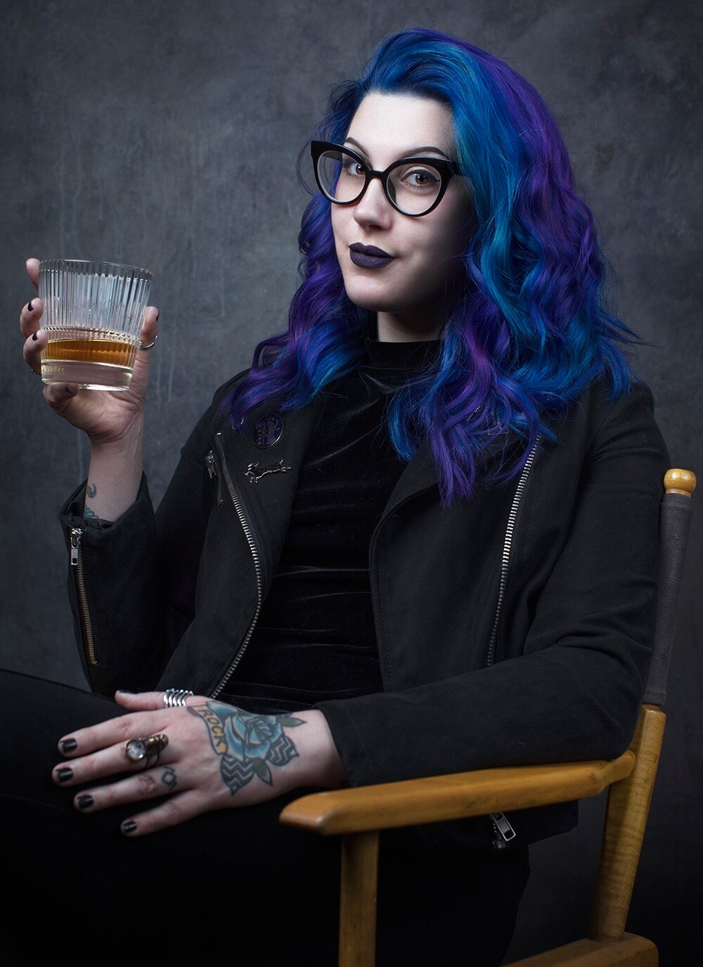 A young woman with blue hair, glasses and a tattoo on her hand holds a drink.