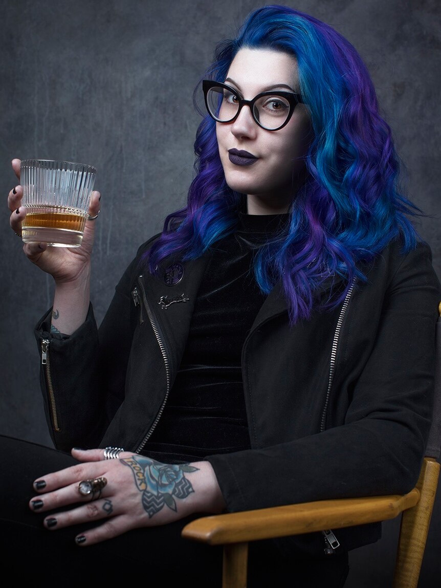 A young woman with blue hair, glasses and a tattoo on her hand holds a drink.
