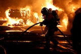 A firefighter holds a hose as a car is alight, covered in bright orange flames, in the background.