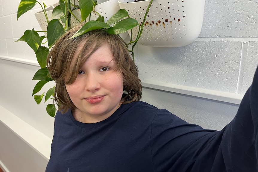 Gus has medium length blonde hair and wears a navy long sleeved shirt. He has a plant box behind him and leaves on his head.