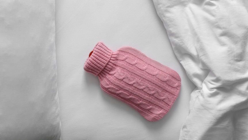 A hot water bottle in a pink knitted cover lies on white sheets in a bed.