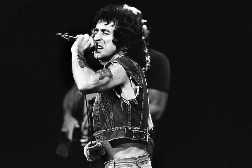 black and white photo of bon scott holding a microphone intensely singing on stage
