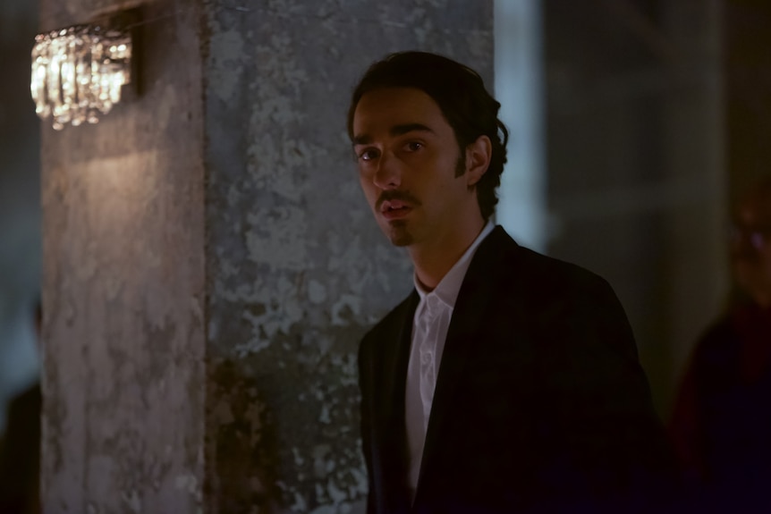 A 20-something man with dark, slicked-back hair and a goatee looks curious. He's wearing a suit and standing in a dark room.