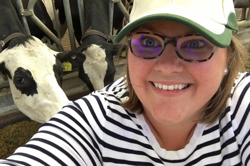 A woman wearing glasses looks at the camera as cows are seen in a milking pen behind.