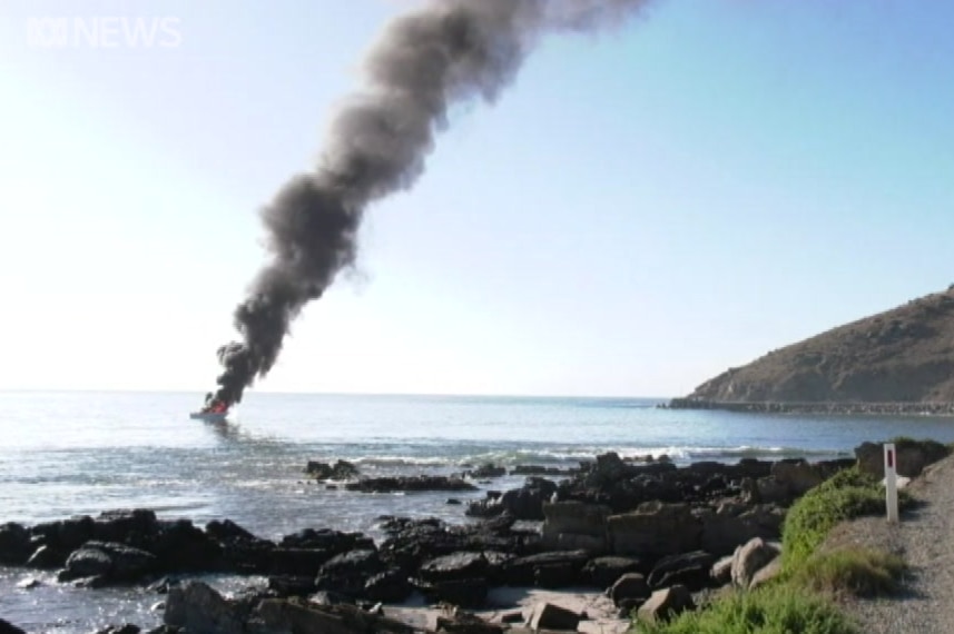 Smoke billowing from a boat in the ocean next to rocks and land