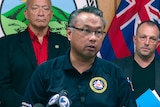 A man wearing glasses and a polo shirt with the Maui emergency service agency logo speaks at a press conference