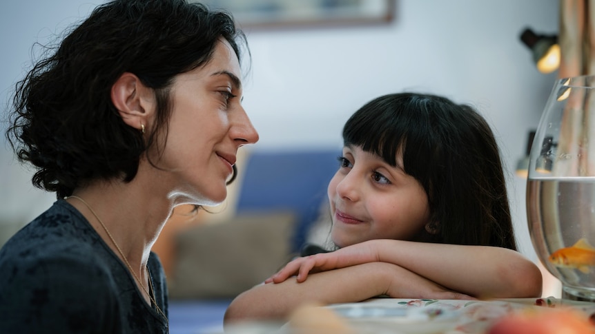 A girl with her arms rested on a table next to a fishbowl, looks up smiling at her mother, who looks at her lovingly.