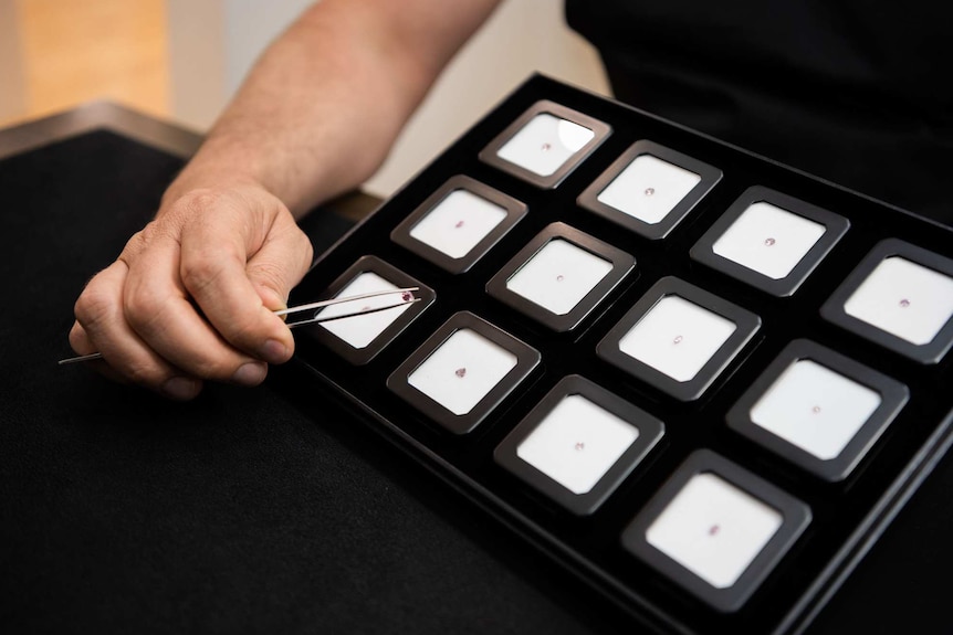 A close-up shot of a man's hands using a pair of tweezers to hold a pink diamond above a tray featuring other diamonds.