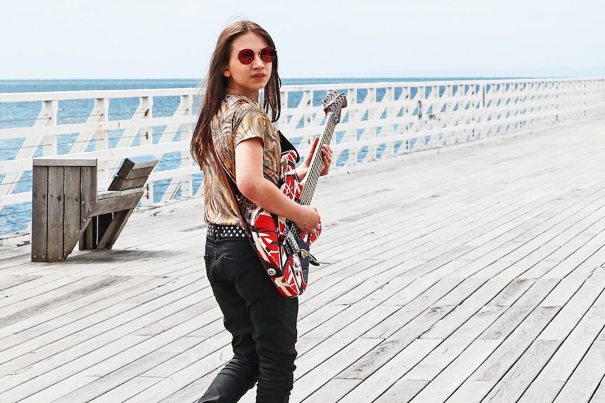 A girl with hair past her shoulders hold an electric guitar on a jetty amidst the sea breeze.