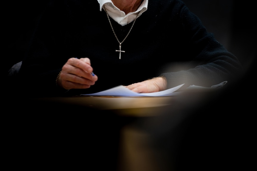 A close up shot of the hands of a person, who is wearing a crucifix, hovering over papers. 