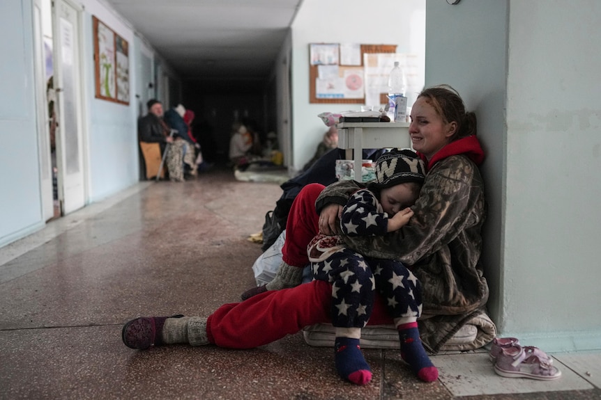 A woman crying sits on the floor of a hospital corridor, hugging a child. Others are seen sitting in the corridor behing them.