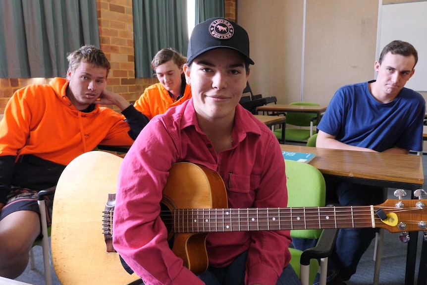 A young woman in a pink shirt and cap holding a guitar with three young men in background.