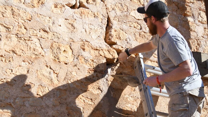 A man with a beard and hat stands on a ladder doing stonework.