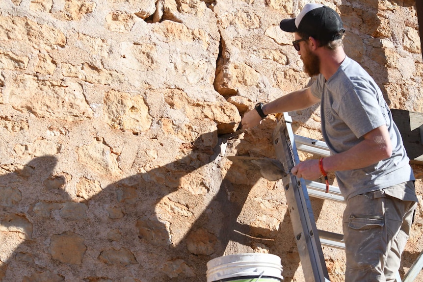 A man with a beard and hat stands on a ladder doing stonework.