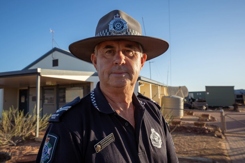 A police officer looking seriously at camera wearing hat and stands in front of a house in outback setting.