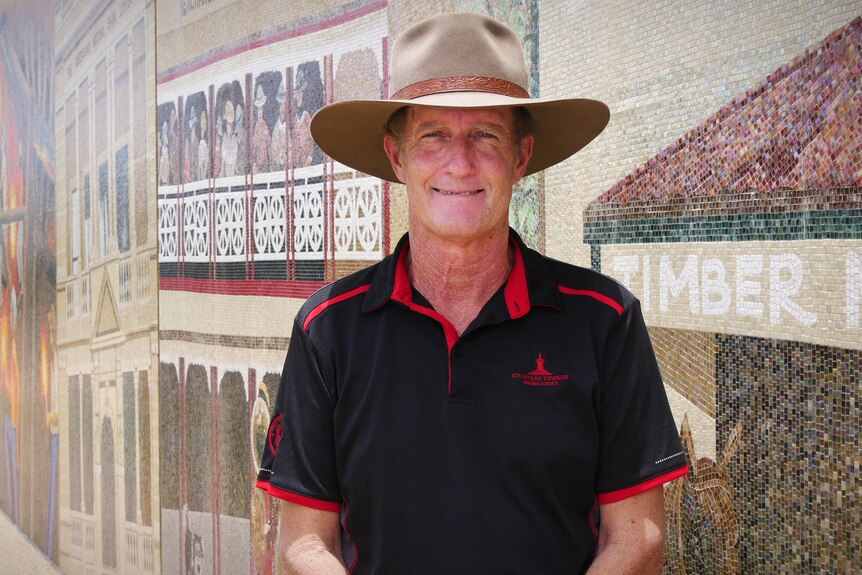 Hombre sonriente con sombrero, camiseta roja y negra, frente a una pared de mosaico, pared cubierta de carteles.  La palabra madera es visible.