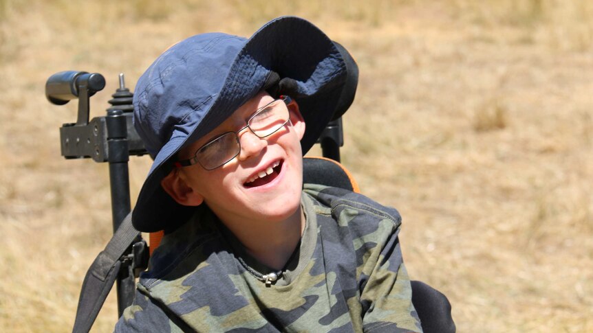 Lucas smiles wide in his wheelchair, the sun on his face, grass around him.