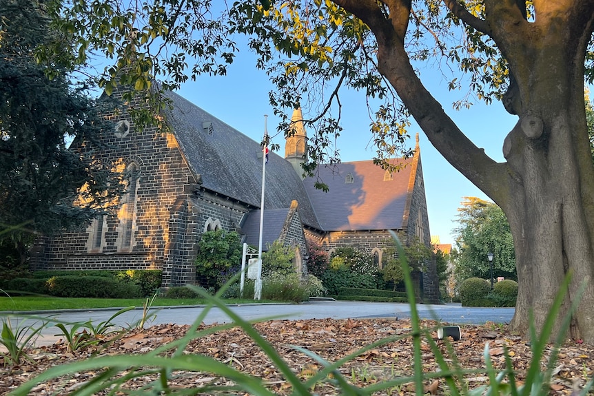 A bluestone church building sits behind a large tree and garden in late afternoon light.