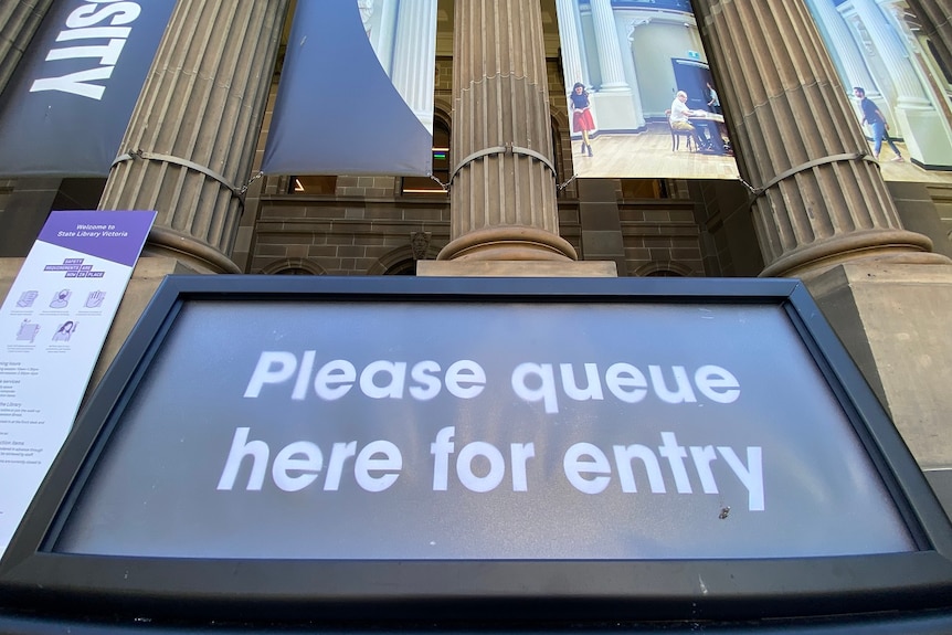 A sign reads 'Please queue here for entry' outside the State Library Victoria.