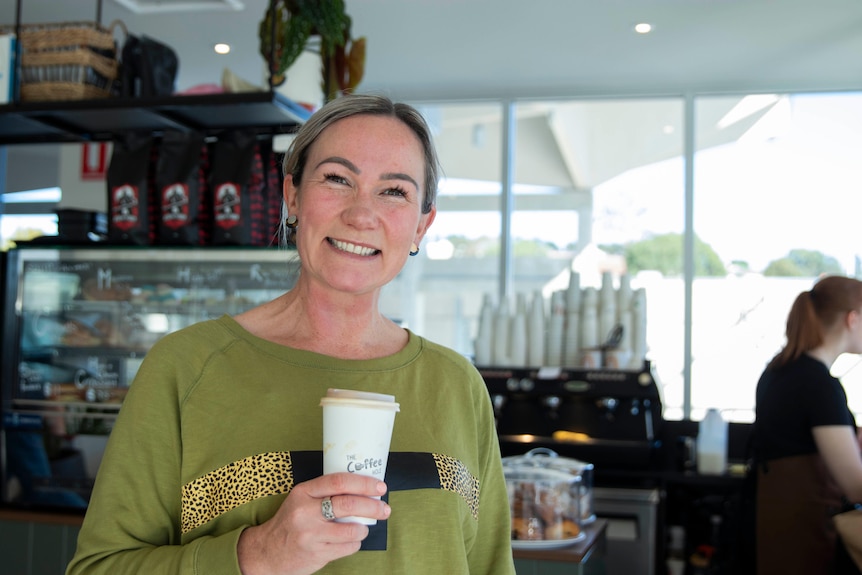 A woman holds a takeaway coffee and smiles.