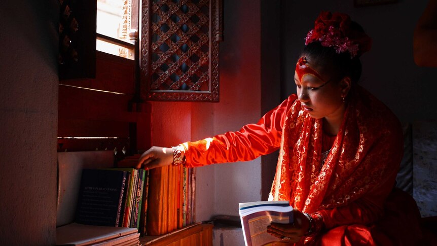 A girl in elaborate traditional dress reaches for a book from a shelf