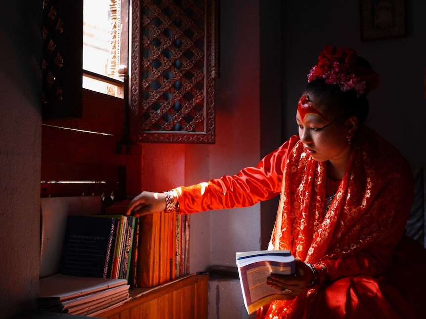 A girl in elaborate traditional dress reaches for a book from a shelf