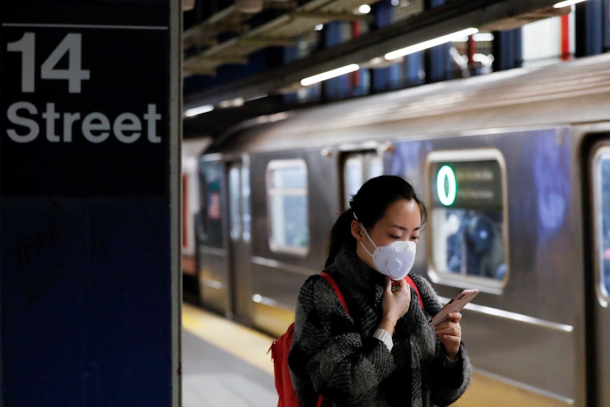 A woman wearing a face mask on a subway platform looks at her mobile phone on a subway.