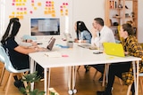 A group of people sit at a table looking at a whiteboard with pink and orange sticky notes on it.