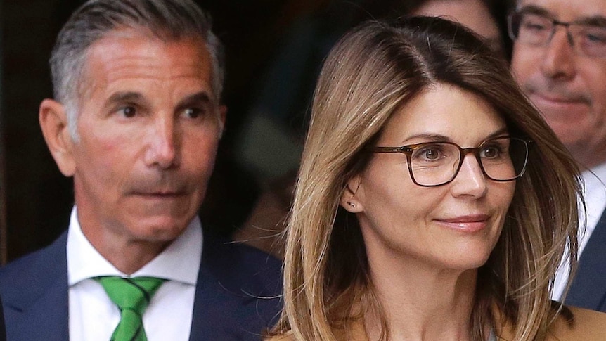 A middle-aged woman wearing glasses looks away from camera as two men in suits stand behind her