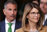 A middle-aged woman wearing glasses looks away from camera as two men in suits stand behind her