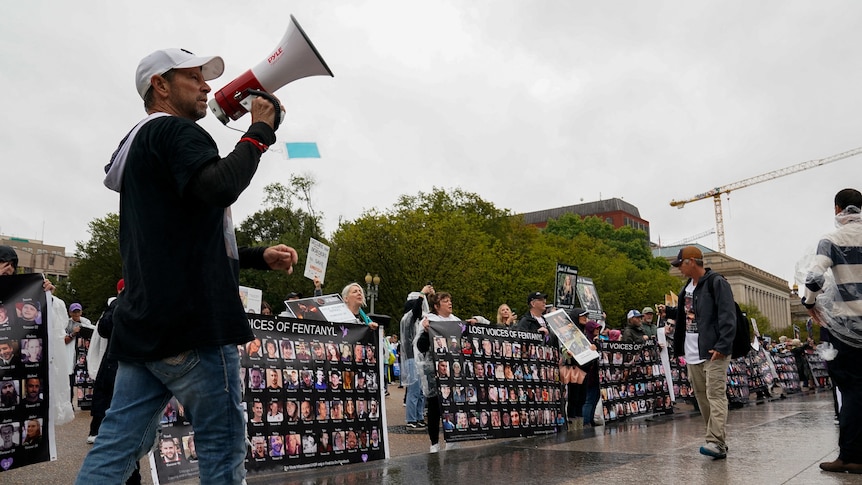 A man holds a megaphone while at a rally, walking past giant posters that contain faces of people who've died from fentanyl