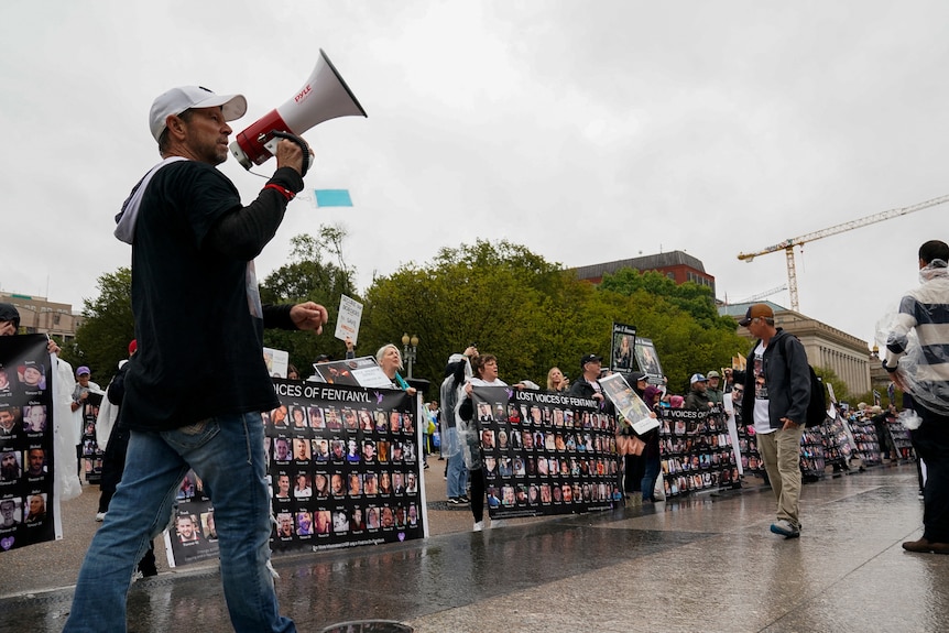 A man holds a megaphone while at a rally, walking past giant posters that contain faces of people who've died from fentanyl