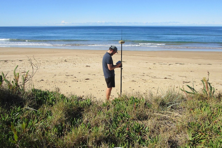 A man with a measuring stick stands on a beach with grass in the foreground and the ocean in the background.