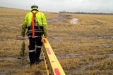 A Western Power worker drags a ladder through a muddy field wearing protective clothing and a helmet and carrying rope.