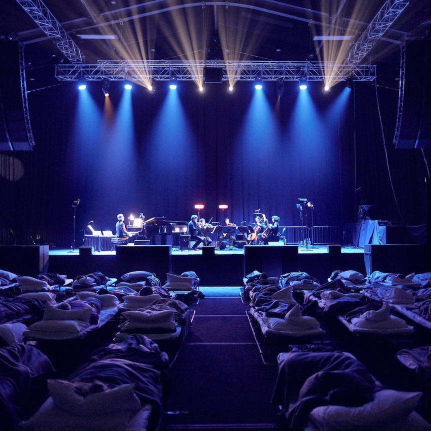 An audience in camp beds listen as musicians on stage perform.