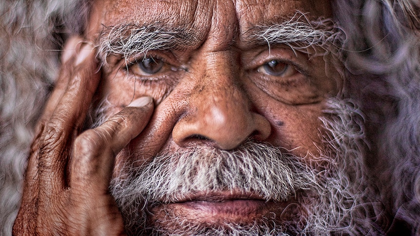 A close up of Uncle Jack Charles holding his hand to face and stares intently at the camera.