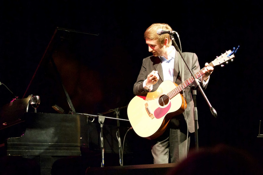 A man in a grey suit and white shirt stands at a microphone on stage with a guitar.