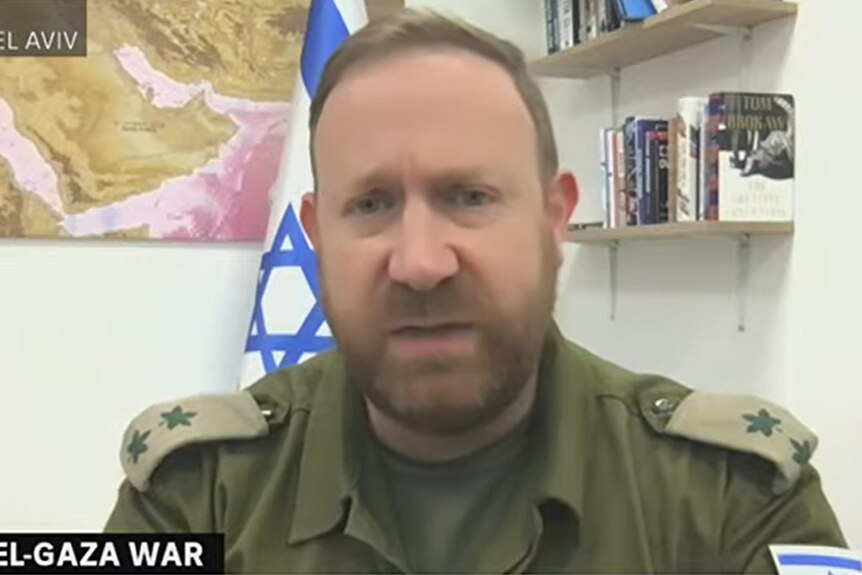 A middle-aged bearded man in an army uniform sits in an office in front of an Israeli flag.