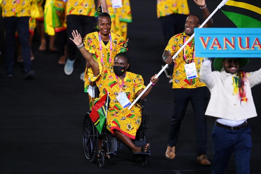 Woman pushed in ability chair holding Vanuatu flag. Wears bright yellow in team of athletes wearing matching outfits.